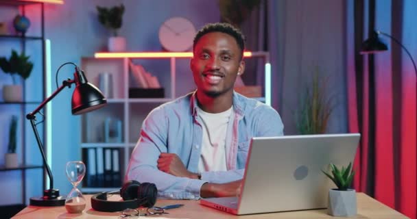 英俊而自信的30岁的非洲裔美国人成功地在他的工作场所的相机上摆出一副迷人的笑容- -晚上用电脑，远程工作的概念 — 图库视频影像