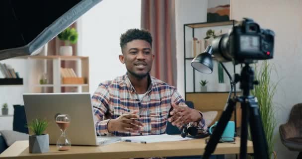 Attrayant sourire jeune homme afro-américain moderne assis devant la caméra à la maison et l'enregistrement de nouvelles vidéos pour ses disciples, blogging et concept d'influence — Video