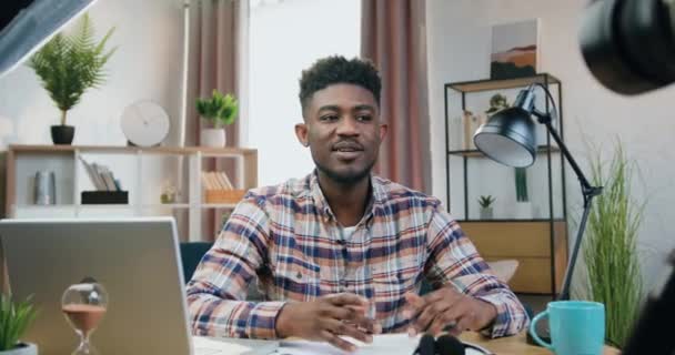 Gut aussehende lächelnde freundliche 25-jährige schwarzhäutige Kerl sitzt vor der Kamera während der Aufnahme von Vlog für Internet-Publikum auf schönen inneren Hintergrund zu Hause — Stockvideo