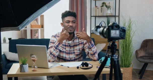 Beau sourire confiant 25 ans homme à la peau noire assis devant la caméra dans le bureau à domicile contemporain lors de l'enregistrement vidéojournal pour les spectateurs sur Internet, concept de blogging — Video