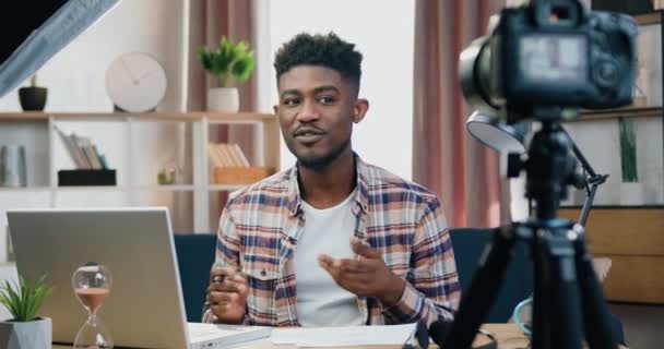 Blogging concept όπου ελκυστικός χαμογελαστός επιτυχημένος νεαρός μαύρος που εργάζεται στον υπολογιστή και καταγράφει βίντεο για τους θεατές του διαδικτύου στην επαγγελματική κάμερα στο σπίτι — Αρχείο Βίντεο