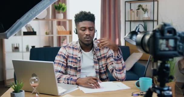 Blogger masculino de piel negra de 25 años de buena pinta y hábil en auriculares sentados frente a la cámara en la oficina en el hogar durante la grabación de un nuevo vlog para su audiencia — Vídeos de Stock