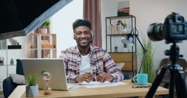 Attraktiver lächelnder erfolgreicher junger schwarzhäutiger Mann mit Kopfhörer und Mikrofon sitzt vor der Kamera in einer Pause zwischen der Aufnahme von Vlog für sein Publikum — Stockvideo