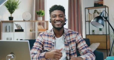 Gülümseyen mutlu, 25 yaşında, kulaklıklı, mikrofonlu, merkez büroda kameranın önünde oturan ve baş parmağını kaldıran Afro-Amerikan adam.