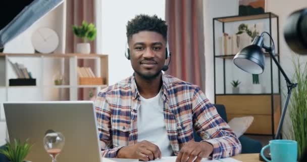 Jóképű mosolygós elégedett fiatal fekete bőrű férfi fülhallgatóval ül a munkahelyén az irodában, és jelképet mutat a kamerába. — Stock videók