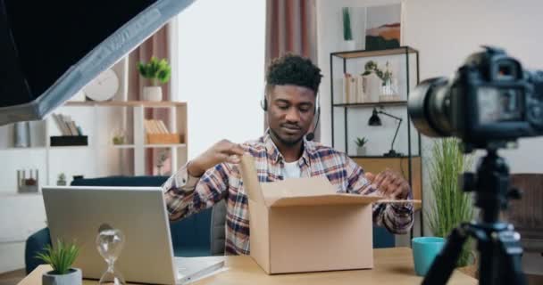 Probablemente sorprendido moderno negro de piel joven blogger masculino sentado frente a la videocamera y la grabación de proceso de desembalaje en el hermoso fondo de interier casa — Vídeos de Stock