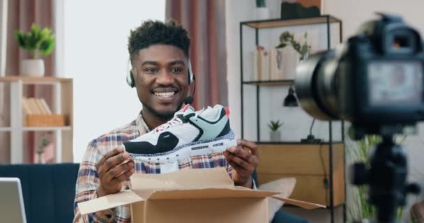 Lidská reakce na koncept nákupu, kde atraktivní šťastný spokojený mladý černoch nahrávání odezvy na kameru pro internetové publikum při vybalení balíčku s tenisky — Stock video