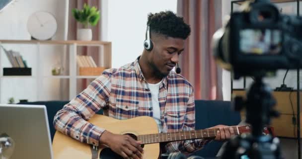 Atractivo chico de piel negra amigable positivo en auriculares inalámbricos grabando su juego de guitarra en videocamera para la audiencia de Internet de su canal de música — Vídeos de Stock