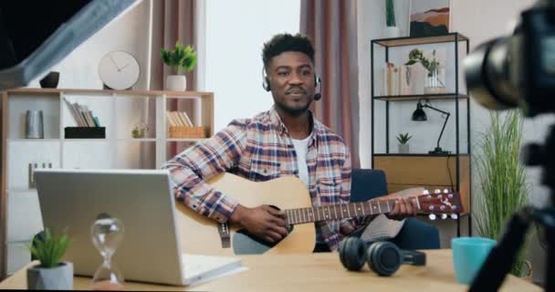 Bonito alegre despreocupado jovem negro de pele cara em fones de ouvido sentado na frente da câmera em casa e gravação de videoblog para seus seguidores de mídia social em guitarra jogar tema — Vídeo de Stock