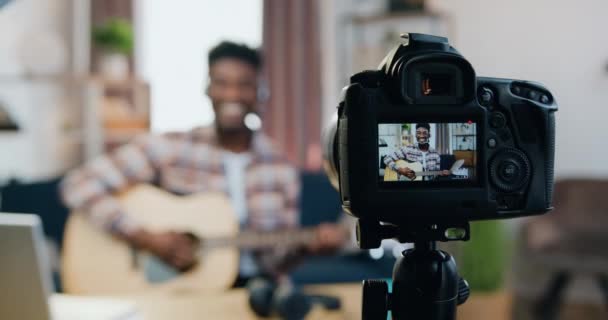 Vue à travers l'objectif de la caméra où sympathique sourire heureux jeune guitariste homme à la peau noire profiter de son jeu à la guitare lors de l'enregistrement d'une nouvelle vidéo pour sa chaîne de musique en utilisant vidéocamera — Video