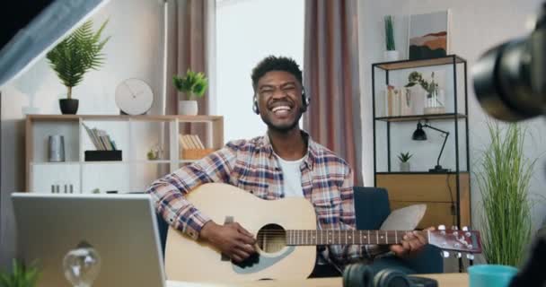 Привабливий сміх безтурботний сучасний молодий чорношкірий хлопець в навушниках тримає гітару в руках і дивиться в камеру з задоволеним виразом обличчя — стокове відео