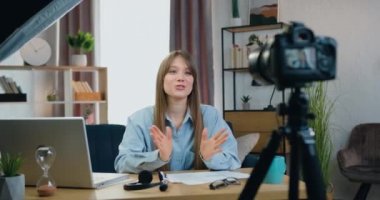 Blog konsepti, güzel, gülümseyen, uzun saçlı, uzun saçlı genç bir kadının evde kameranın önünde oturup izleyicileri için canlı yayında konuştuğu.