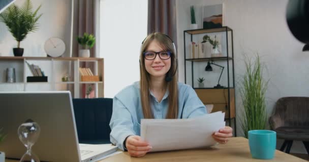 Jolie jeune fille souriante aux cheveux longs dans des écouteurs assis devant la caméra avant le début du flux en ligne avec des documents préparés sur son travail — Video