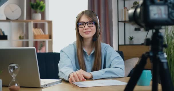 기분좋게 기분좋게 성공 한 젊은 소녀가 안경 과 이어폰을 끼고 집 사무실에 앉아서 카메라 앞에서 앞을 보고 느린 동작을 하며 바쁘게 움직 인다 — 비디오
