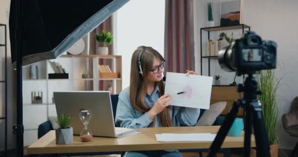 Concept d'apprentissage à distance où attrayant confiant jeune fille qualifiée dans les écouteurs assis devant la caméra dans le bureau à domicile et la tenue d'une conférence en ligne expliquant rapport avec des graphiques — Video