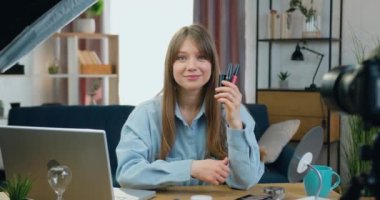 Gülümseyen, kendine güvenen genç bir bayan güzellik blogcusu kozmetik ürünlerini bir elinde tutuyor ve ev stüdyosundaki kameraya el sallıyor.