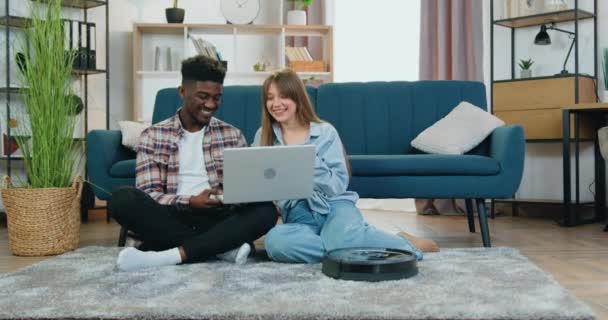 Conceito de função casa inteligente onde atraente satisfeito cara multirracial e menina sentada no chão e usando laptop enquanto robô aspirador de pó tapete de limpeza — Vídeo de Stock