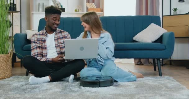 Modern robot porszívó tisztítás bolyhos szőnyeg közel szerethető pozitív mosolygós fiatal többnemzetiségű pár, amely érdekes videót néz laptop a padlón — Stock videók