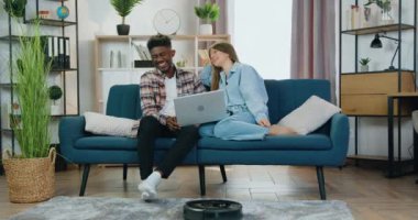 Modern ev konsepti, siyah robot elektrikli süpürge temizlik halısını temizlerken aynı zamanda çekici gülümseyen karışık ırk arkadaşlarının kanepede oturup dizüstü bilgisayarda video izlerken sohbet etmesi.