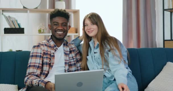 Bello felice sorridente giovane due ragazzo multietnico e ragazza seduta sul divano a casa durante la revisione interessante programma sul computer portatile e in posa sulla macchina fotografica con volti soddisfatti — Video Stock