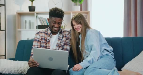 Aantrekkelijke vrolijke jonge multiraciale twee mannelijke en vrouwelijke vrienden zitten samen op comfortabele bank thuis en herzien grappige video 's op de computer — Stockvideo