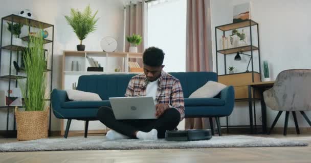 Концепція розумного будинку, де добре виглядає емоційний 20-річний афроамериканський хлопець сидить на підлозі біля дивана вдома і працює на ноутбуці під час автоматичної чистки пилососа — стокове відео