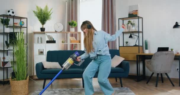 Schöne positive lustige junge moderne Mädchen in Kopfhörern Staubsauger Teppich mit Staubsauger zu Hause und genießen Lieblingssongs, Housekeeping-Konzept — Stockvideo