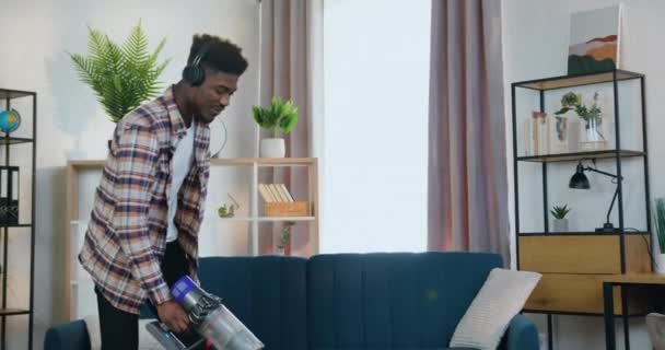 Beau sourire heureux énergique jeune homme à la peau noire tapis hoovering avec aspirateur et écouter de la musique dansante dans les écouteurs, concept de ménage — Video