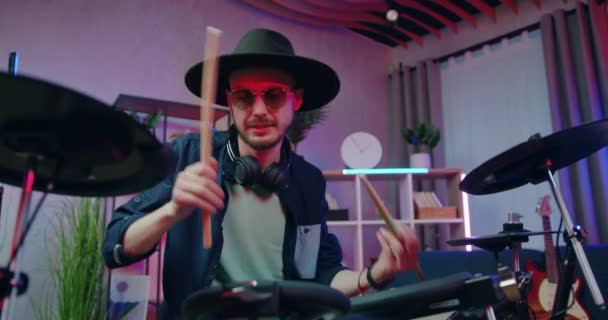 Музична концепція, де добре виглядає емоційно щасливий усміхнений молодий барабанщик у чорному капелюсі грає на барабанах увечері в домашній студії, вид спереду — стокове відео