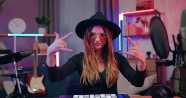 Ελκυστική χαμογελαστή επιτυχημένη νεαρή κομψή καλλιτέχνιδα DJ με καπέλο και γυαλιά κάθεται στο DJ σετ στο σπίτι στούντιο το βράδυ και gesturing διαφορετικές πινακίδες στην κάμερα — Αρχείο Βίντεο