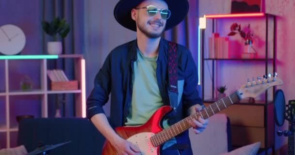 Vonzó pozitív ügyes, fiatal, stílusos, szakállas fickó kalapban és szemüvegben, aki az előadásra készül, és este a gitáron próbál a hazai stúdióban. — Stock videók
