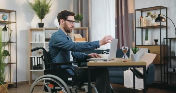 Vonzó magabiztos ügyes, 40 éves szakállas mozgássérült férfi tolószékben dolgozik laptop otthoni irodában nappal — Stock videók