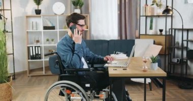 Çekici, gülümseyen, sakallı, tekerlekli sandalyede sakallı bir adam ev bilgisayarında çalışıyor ve aynı zamanda iş arkadaşı ya da müşterisiyle akıllı telefondan konuşuyor.