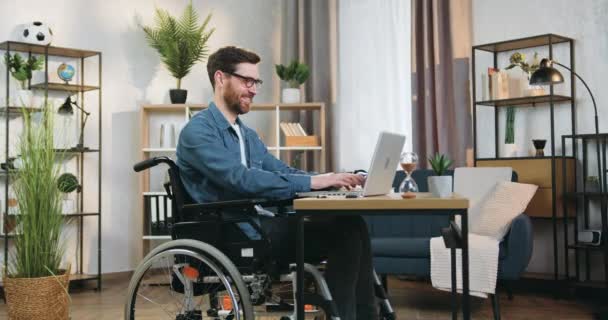 英俊、积极、自信的成年人坐在轮椅上，坐在笔记本电脑前，在远离总部的地方为商业项目工作 — 图库视频影像