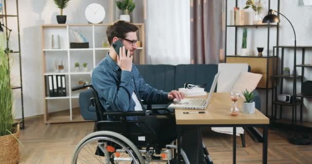 Atractivo sonriente adulto barbudo hombre discapacitado en silla de ruedas trabajando en el ordenador portátil en la oficina en casa y hablando simultáneamente en el teléfono inteligente con su colega o cliente — Vídeo de stock