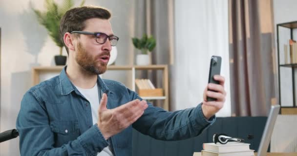 Hezký jistý pozitivní dospělý vousatý handicapovaný muž v brýlích sedí na vozíku a mluví na videokallu pomocí smartphonu v domácí kanceláři — Stock video