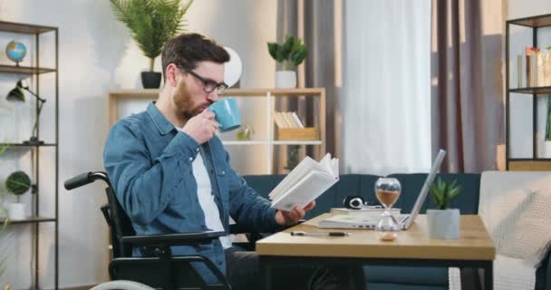 Tiltrekkende fornøyd, avslappet, voksen, skjeggete mann i briller som sitter i rullestol og leser bok samtidig som han drikker te hjemme – stockvideo