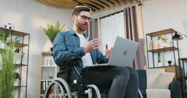 Primer plano de simpático sonriente hombre con discapacidad barbuda de 40 años en silla de ruedas después del acento que sostiene el ordenador de rodillas durante el videochat con un amigo o pariente en casa — Vídeo de stock