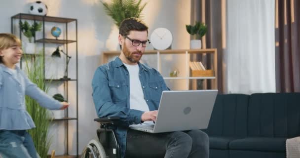 Guapo positivo exitoso adulto barbudo hombre discapacitado en gafas en silla de ruedas trabajando en el ordenador portátil, mientras que la hija adolescente bastante alegre corriendo hacia él y abrazándose por el cuello — Vídeos de Stock