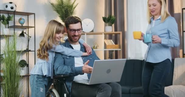 Family idyll where beautiful adult blondie giving two cups of tea to her cute smiling teenage daughter and handsome bearded disabled husband in wheelchair which together applying laptop at home — Stock Video