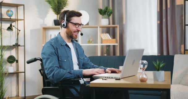 Hombre barbudo adulto hábil y seguro de buena apariencia en auriculares sentados en silla de ruedas delante de la computadora portátil durante la reunión de video en línea con socios de forma remota desde la oficina en el hogar — Vídeo de stock