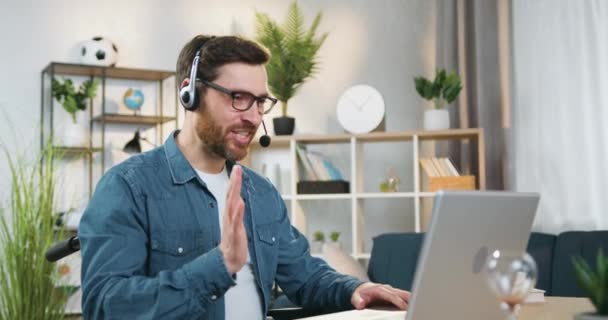 Concepto de trabajo remoto donde agradable hombre barbudo profesional positivo adulto en auriculares sentados en silla de ruedas después de un trauma en casa durante la videoconferencia en línea con clientes o compañeros de trabajo — Vídeos de Stock