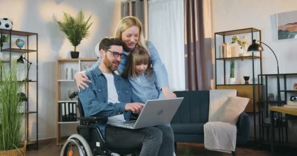 Attrayant sourire adulte handicapé en fauteuil roulant application ordinateur portable avec sa fille mignonne de 10 ans et jolie femme joyeuse à la maison — Video