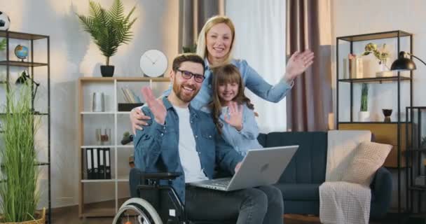 Portret van goed uitziende gelukkig lachende moderne familie van tienermeisje, mooie vrouw en bebaarde gehandicapte man in rolstoel die poseren op camera met zwaaiende handen binnen — Stockvideo