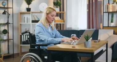 Kendine güvenen, başarılı, açık renk saçlı, tekerlekli sandalyedeki engelli kadın güzel dekore edilmiş ev ofisinde dizüstü bilgisayarda çalışıyor, ağır çekimde.