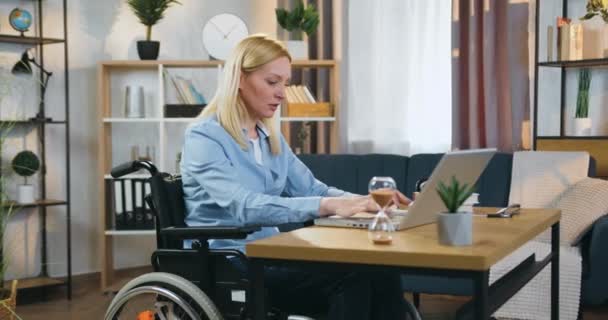 Mooie zelfverzekerde succesvolle volwassen licht-harige gehandicapte vrouw in rolstoel werken op laptop in prachtig ingerichte kantoor thuis, slow motion — Stockvideo