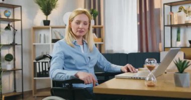 Güzel görünüşlü, kendine güvenen, 40 yaşında, açık renk saçlı, sıradan elbiseli, tekerlekli sandalyede oturan ve kameraya baş parmağını gösteren bir kadın.