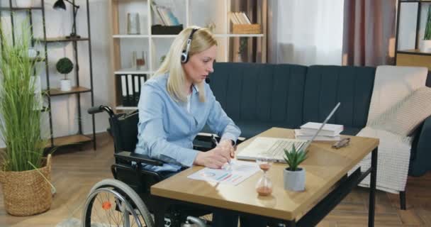 Очаровательная серьезная аттантивная взрослая работница, временно прикованная к инвалидному креслу после несчастного случая, сидит перед ноутбуком в наушниках во время видеочата и делает заметки в отчете — стоковое видео