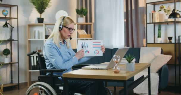 Atraktivní moudrý úspěšný dospělý hendikepovaná žena ve sluchátkách sedí na kolečkovém křesle a pořádá on-line setkání s kolegy a vysvětluje zprávu s grafy — Stock video