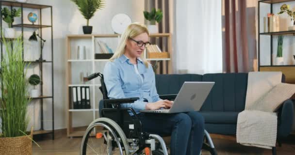 Docela veselý 10-leté dívka běží ke své krásné zkušené světlovlasé matky, která sedí na invalidním vozíku po nehodě a pracuje na počítači doma — Stock video